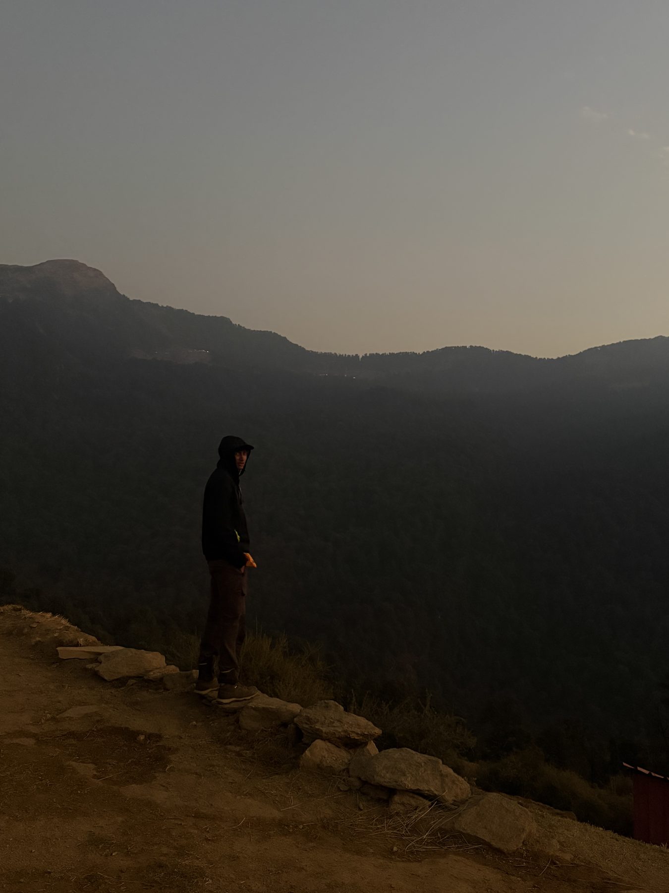 Hooded figure on a rocky ledge looking back over hazy ridges